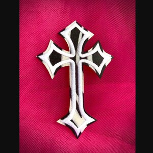 May include: A decorative cross patch with a white border and a black and white patterned center. The cross is set against a bright pink background, creating a striking contrast. The patch is likely for clothing or accessories.