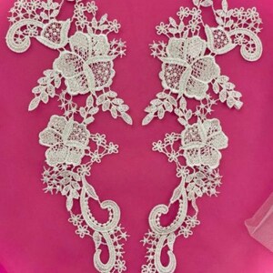May include: Two white lace appliques with floral and leaf designs. The appliques are symmetrical, featuring large flowers, delicate leaves, and intricate scrollwork. The background is a vibrant pink.
