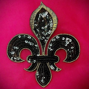 May include: A black fleur-de-lis emblem with gold edging and sequins, set against a bright pink backdrop. The emblem's design features three stylized petals and a central vertical element, all embellished with shimmering sequins.