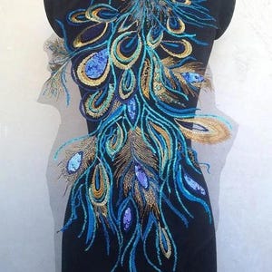 Puede incluir: Tela negra con un diseño de pluma de pavo real bordada. El diseño presenta hilos azules, dorados y turquesas, creando un patrón detallado y ornamentado. El diseño de pluma de pavo real es un motivo popular para ropa y accesorios.