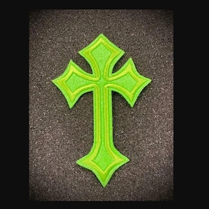 May include: A neon green embroidered cross patch with a lighter green outline. The cross is centered on a dark, textured background. The patch has a detailed, layered design, suitable for clothing or accessories.