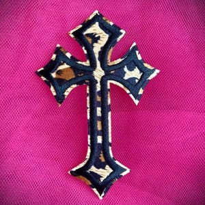 May include: A decorative cross-shaped patch with a leopard print design and navy blue trim. The patch is centered on a bright pink textured background.