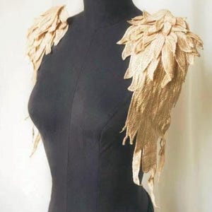 May include: A gold-colored shoulder piece with a leaf-like design. The shoulder piece is made of a textured material and is displayed on a black mannequin. The leaves cascade down the arms.