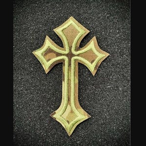 May include: A decorative cross patch with a camouflage pattern and a light green border. The cross has a detailed design with pointed edges and a textured appearance. The patch is likely for clothing or accessories.