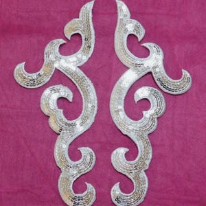 May include: Two silver sequined appliques with an ornate, swirling design. The appliques are symmetrical, with a white border and a pattern of silver sequins. The background is a solid magenta color.