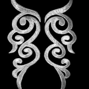 Puede incluir: Dos apliques bordados plateados simétricos con un diseño ornamental y arremolinado. Cada aplique presenta una forma curva, similar a un cuerno, en la parte superior y una serie de rizos y bucles interconectados. Los apliques están sobre un fondo negro sólido.