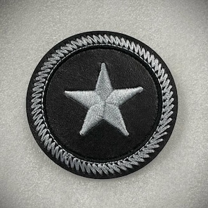 Peut inclure: Un écusson rond noir avec une étoile brodée argentée au centre. L'étoile est à cinq branches. L'écusson a une bordure brodée argentée. Le fond est gris clair.