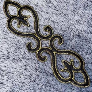 Puede incluir: Aplicación bordada en negro y dorado con un diseño simétrico y ornamentado. La aplicación presenta patrones arremolinados y acentos florales, con un contorno dorado y un interior negro. El elemento decorativo se encuentra sobre un fondo gris texturizado.