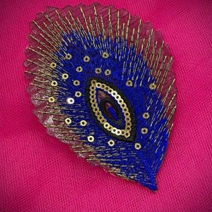 May include: A decorative, leaf-shaped embellishment with a vibrant blue center, outlined with gold beads and sequins. The edges are a sheer, reddish-brown fabric. The background is a bright pink.