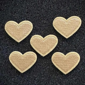 May include: Five embroidered heart patches. Each heart is a light tan color with a cream-colored border. The patches are arranged in a scattered pattern against a dark, textured background. These patches are ideal for adding a decorative touch to clothing or accessories.