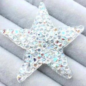 May include: A clear, five-pointed star-shaped embellishment, encrusted with numerous iridescent rhinestones. The rhinestones reflect a spectrum of colors, creating a sparkling effect. The star is set against a soft, gray background.