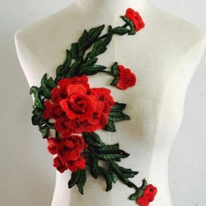 May include: A vibrant red rose applique with detailed embroidery, featuring lush green leaves. The floral design is displayed on a mannequin, showcasing the intricate craftsmanship of the fabric embellishment. This decorative patch is ideal for clothing and craft projects.