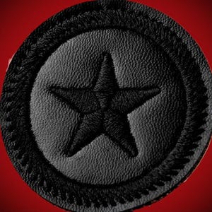 Puede incluir: Un parche de cuero negro con un diseño de estrella. La estrella está bordada en negro y centrada dentro de un parche circular. El borde del parche tiene un borde decorativo cosido. El fondo es rojo liso.