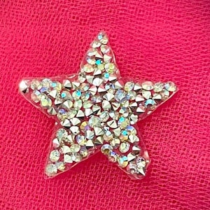 May include: A star-shaped embellishment covered in iridescent rhinestones. The star is set against a textured, bright pink background. The rhinestones reflect multiple colors, creating a sparkling effect.