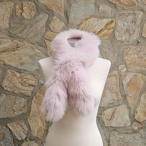May include: A light pink fur scarf displayed on a mannequin. The scarf is made of soft, fluffy fur and has a loop at the top for wearing around the neck. The background is a stone wall.