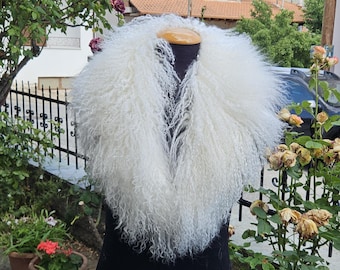 May include: A fluffy, white fur collar displayed on a black mannequin. The collar has a dense, curly texture and is designed to be worn around the neck. The background features a garden with flowers and a view of a town.