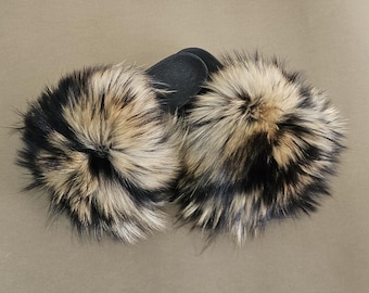 May include: A pair of fluffy, faux fur pom-poms in shades of black, tan, and cream. The pom-poms are attached to a black leather base. The fur is dense and textured, creating a luxurious appearance.