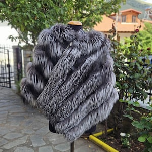 May include: A silver fox fur jacket on a mannequin. The jacket has a layered design with long, flowing fur. The fur has a mix of silver and dark grey tones, creating a textured appearance. A gold zipper is visible.