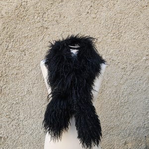 Puede incluir: Bufanda negra y esponjosa exhibida en un maniquí. La bufanda tiene una apariencia texturizada y peluda, con hebras largas y fluidas. El fondo es una pared texturizada de color marrón claro.