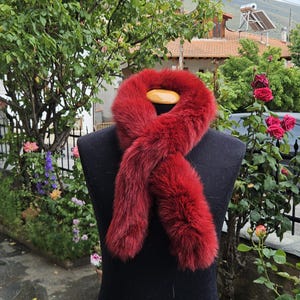May include: A vibrant red fur scarf displayed on a black mannequin. The scarf is made of soft, plush fur and is draped in a loop. The background features lush greenery and a glimpse of a building.