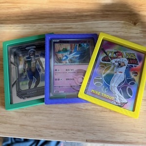 May include: Three rectangular trading card display frames in green, purple, and yellow. The green frame holds a football player card, the purple frame contains a purple Pokémon card with a blue creature, and the yellow frame holds a Mike Trout baseball card.
