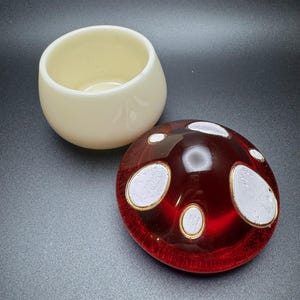 May include: A cream-colored, rounded ceramic cup next to a red glass paperweight. The paperweight has white and gold accents. The objects are on a dark gray surface.
