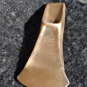 Peut inclure: Une tête de hache en métal de couleur bronze dotée d'un tranchant large et incurvé ainsi que d'une douille rectangulaire sur le dessus pour le manche. La surface présente une finition métallique lisse avec des variations de ton et de texture.