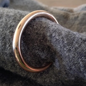 Op de afbeelding: Een glanzende, koperkleurige ring omsluit een deel van grijze, getextureerde stof. De ring is glad en reflecterend, wat contrasteert met de ruwe, wollen textuur van de stof. De afbeelding is een close-up, die de details benadrukt.