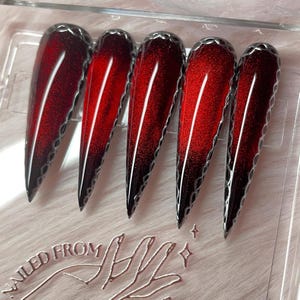 May include: Five long, pointed artificial nails with a gradient design, transitioning from a deep red to black. Each nail features a silver, scalloped edge and a central white stripe. The image includes the text "NAILED FROM".