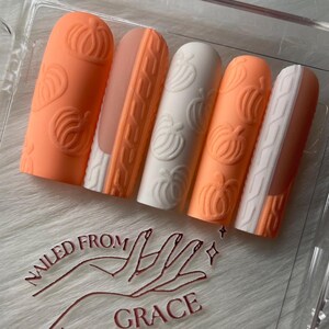 May include: A set of five press-on nails in a clear plastic case. The nails feature a pumpkin theme with orange and white colors. Some nails have a cable knit design, while others have pumpkin carvings. The text "NAILED FROM GRACE" is visible.