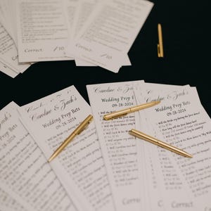 May include: Wedding prop bet cards with gold pens. The cards are white with black text, featuring the names "Caroline & Zach" and the date "09-28-2024". The pens are gold and lie across the cards.