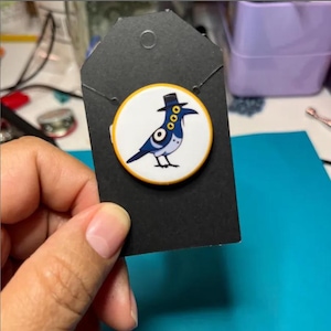 Puede incluir: Un botón redondo con un pájaro de dibujos animados con un sombrero negro, sobre un fondo blanco con un borde amarillo. El pájaro es azul y blanco, sujeto a una tarjeta negra.