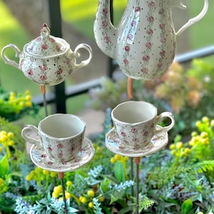 May include: A whimsical garden tea set includes a teapot, a small teapot, and two teacups with saucers. The white ceramic pieces are decorated with red roses and black dots. The set is mounted on metal stakes.