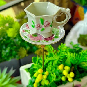 May include: A miniature white teacup and saucer set with pink floral patterns and gold edging. The teacup has an octagonal shape and a heart-shaped handle. The set is displayed above a small planter with green and yellow foliage.