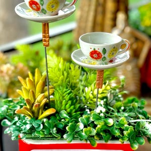 May include: Two decorative teacup and saucer garden stakes. The white ceramic cups and saucers are adorned with red and yellow floral designs. The stakes have a brown, textured appearance and are set in a bed of greenery.