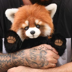 Puede incluir: Un peluche de panda rojo con una cabeza marr&oacute;n rojiza y esponjosa, orejas blancas y patas negras. El juguete tiene una nariz negra y bigotes. El texto "With love Lazienka" es visible en la imagen.