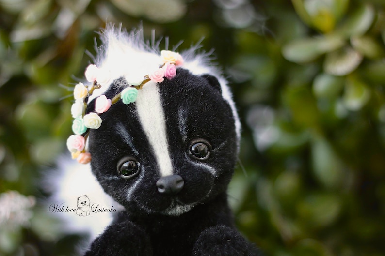 May include: A black and white skunk plush toy with a white stripe down its back. The skunk is wearing a flower crown with pink, white, and yellow flowers. The toy is looking at the camera with big, black eyes.