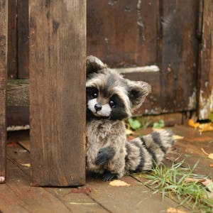 K&ouml;nnte beinhalten: Ein handgefertigtes Pl&uuml;schtier eines Waschb&auml;ren mit einem schwarz-grau gestreiften Schwanz und Fell. Der Waschb&auml;r versteckt sich hinter einem Holzpfosten. Das Spielzeug hat einen niedlichen Ausdruck mit schwarzen Augen und einer wei&szlig;en Schnauze.