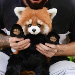 Puede incluir: Un peluche de panda rojo con cabeza marr&oacute;n y blanca, patas negras y cola esponjosa. El peluche est&aacute; siendo sostenido. El texto "With love Lasenka" es visible.