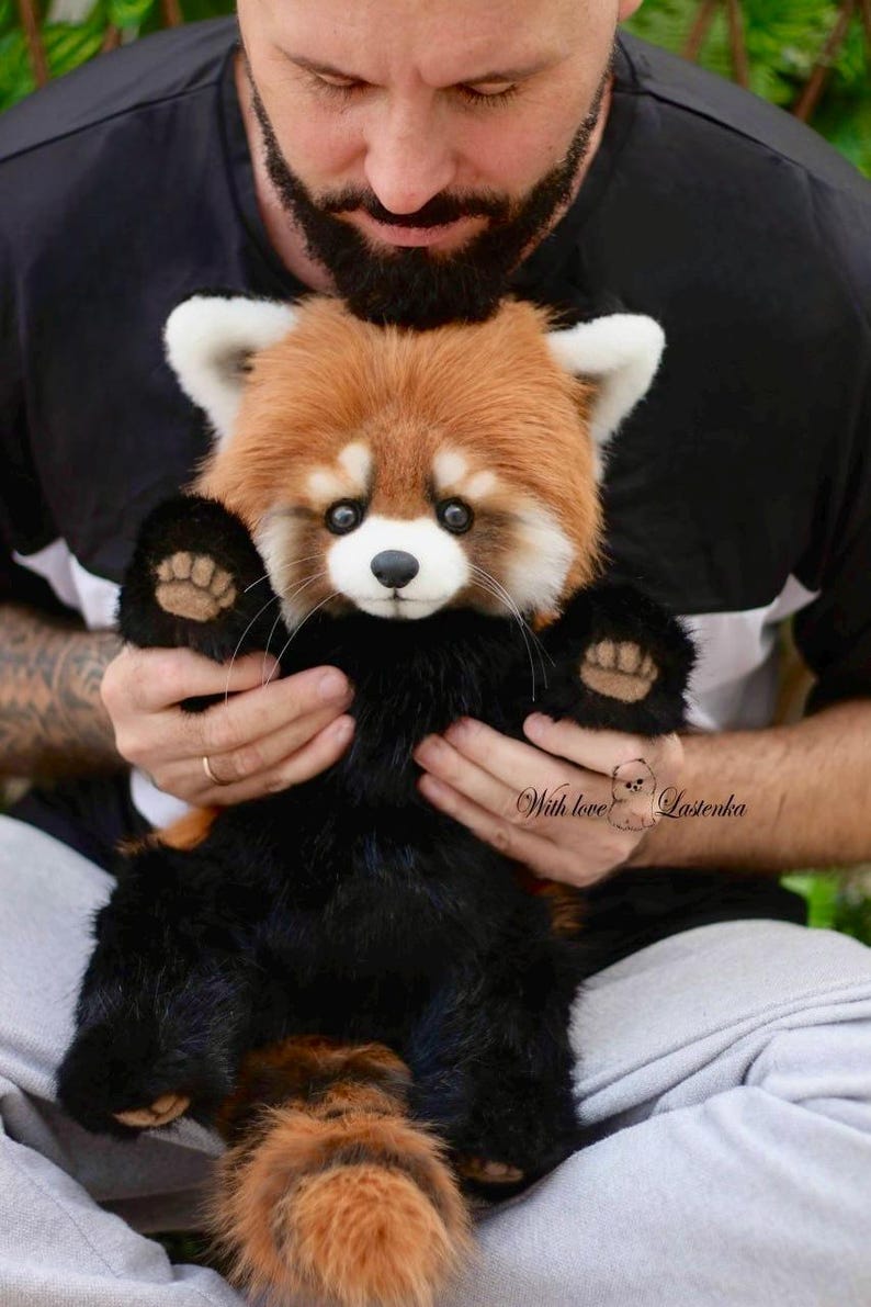 Puede incluir: Un peluche de panda rojo con cabeza marr&oacute;n y blanca, patas negras y cola esponjosa. El peluche est&aacute; siendo sostenido. El texto "With love Lasenka" es visible.
