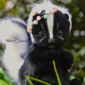 May include: A black and white skunk plush toy wearing a flower crown. The skunk is sitting in a bed of green grass with white flowers.