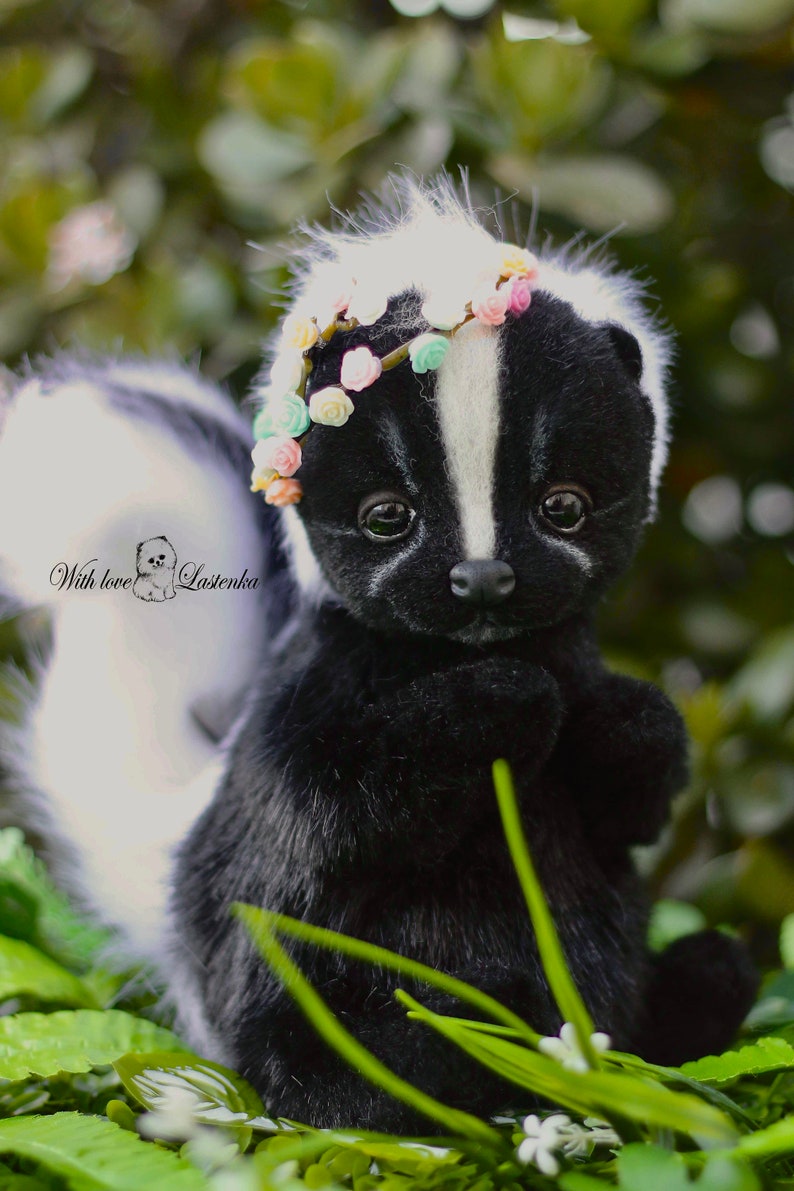 May include: A black and white skunk plush toy wearing a flower crown. The skunk is sitting in a bed of green grass with white flowers.