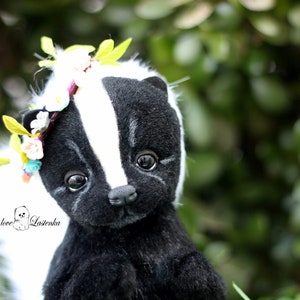 May include: A black and white stuffed skunk with a white tail, wearing a flower crown with green leaves. The skunk has black eyes and a black nose. The background is blurry and green.