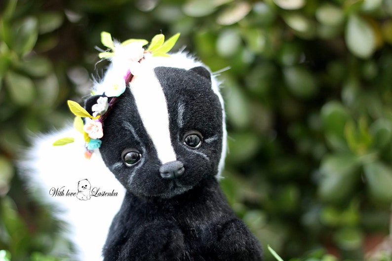 May include: A black and white stuffed skunk with a white tail, wearing a flower crown with green leaves. The skunk has black eyes and a black nose. The background is blurry and green.