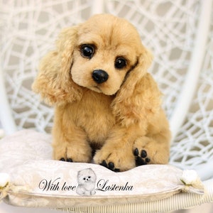 May include: A plush toy of a golden cocker spaniel puppy sitting on a white cushion with the text "With love Lastenka".