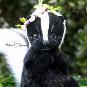 May include: A black and white skunk plush toy wearing a flower crown. The skunk is sitting on a bed of green leaves and white flowers. The text "With love, Lastenka" is visible in the lower left corner of the image.