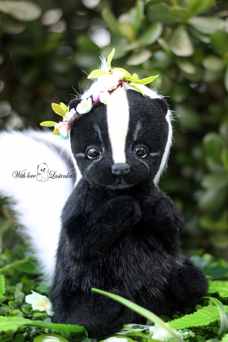 May include: A black and white skunk plush toy wearing a flower crown. The skunk is sitting on a bed of green leaves and white flowers. The text "With love, Lastenka" is visible in the lower left corner of the image.