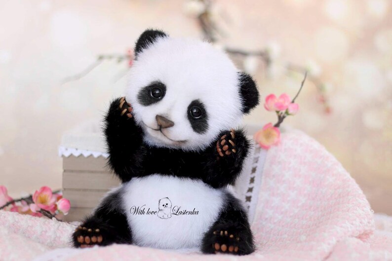May include: A handmade panda bear plush toy with black and white fur, sitting with its arms raised. The panda has a sweet expression and is surrounded by pink flowers and a soft pink blanket. The text "With love" is visible on the panda's belly.