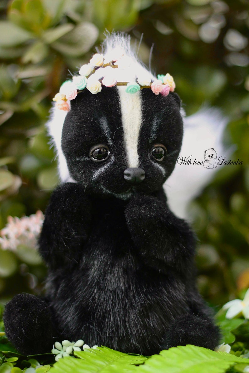 May include: A black and white skunk plush toy wearing a flower crown. The toy has a white stripe down its back and a cute, innocent expression. The toy is sitting on a bed of green leaves.