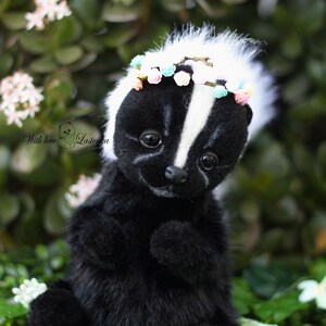 May include: A black and white skunk plush toy wearing a flower crown. The skunk is sitting on a bed of green leaves.